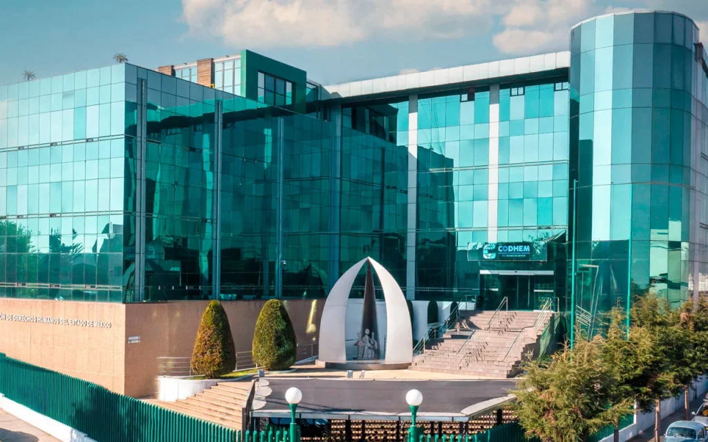 Edificio sede de la CODHEM. Imagen de un edificio moderno con fachada de cristal en tonos azules, acceso principal con escalinatas y barandales, y banderas institucionales y nacionales colocadas al frente.