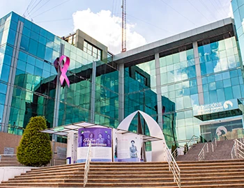 Edificio institucional con fachada de vidrio y escalinatas de acceso al frente. En la parte superior se observa un listón rosa como símbolo de concientización sobre el cáncer de mama. Imagen acompañada del mensaje: “TODAS LAS MUJERES DEBEN TENER ACCESO A LA INFORMACIÓN PARA DETECTAR Y PREVENIR EL CÁNCER DE MAMA: CODHEM”.