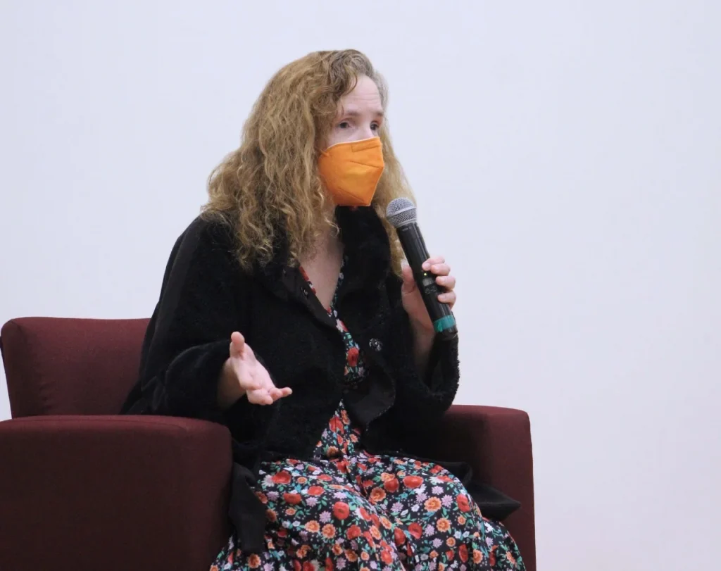 Persona sentada en el escenario, usando cubrebocas y sosteniendo un micrófono, participando en una mesa de diálogo.