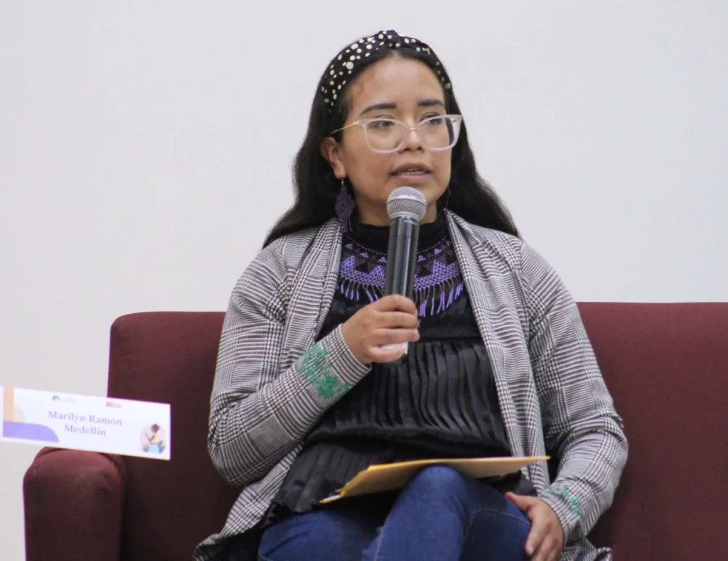 Persona sentada en un sillón, con lentes, hablando al micrófono durante un evento académico o institucional.