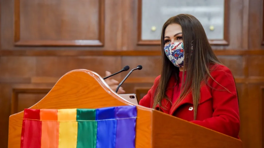 La presidenta de la Comisión de Derechos Humanos del Estado de México (Codhem) Myrna Araceli García Morón participa en una intervención desde un podio con bandera arcoíris. Hizo un exhorto a la LXI Legislatura del Estado de México para reparar la discriminación normativa en la legislación mexiquense sobre el matrimonio igualitario.