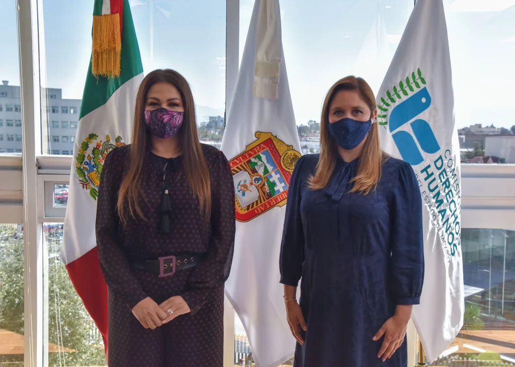 Dos personas posan de pie frente a banderas institucionales durante un encuentro oficial. La presidenta de la Comisión de Derechos Humanos del Estado de México (Codhem) Myrna Araceli García Morón y la presidenta municipal de San Mateo Atenco, Ana Aurora Muñiz Neyra, acordaron diseñar y ejecutar acciones para promover el respeto a la dignidad de las personas del municipio.