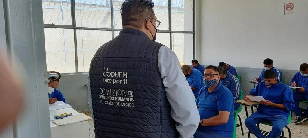 Personal con chaleco institucional de la Codhem dialoga con personas sentadas en un aula dentro de un centro penitenciario. La Comisión de Derechos Humanos del Estado de México (Codhem) participó en la aplicación del Diagnóstico Nacional de Supervisión Penitenciaria 2022.