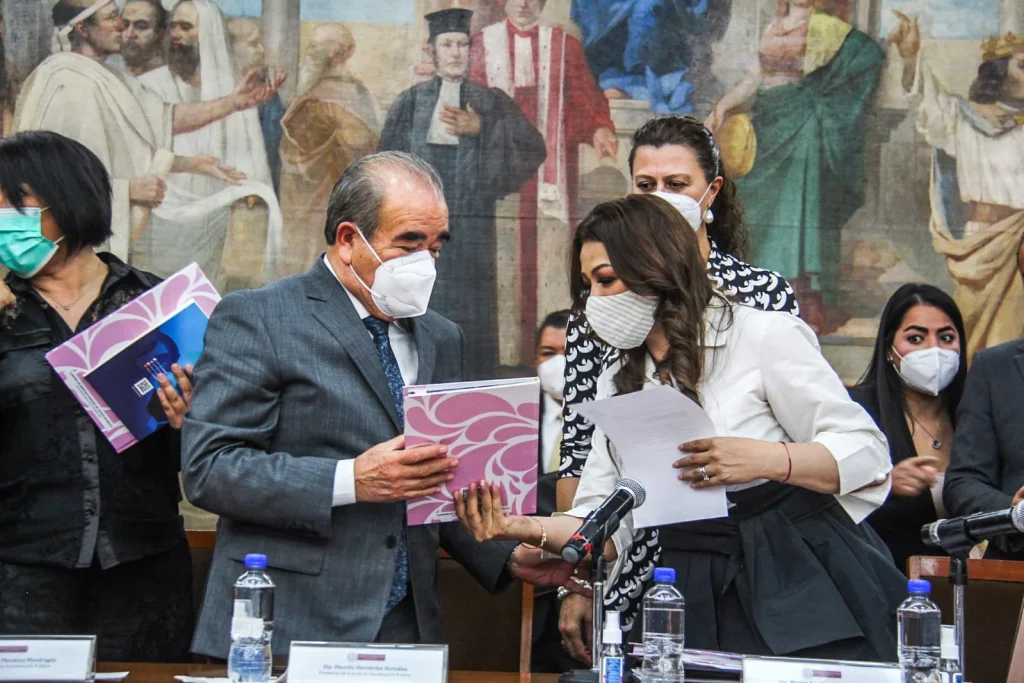 Personas en un acto oficial intercambian documentos en un recinto legislativo. La presidenta de la CODHEM, Myrna Araceli García Morón, entregó su Informe de Actividades a la LXI Legislatura local.