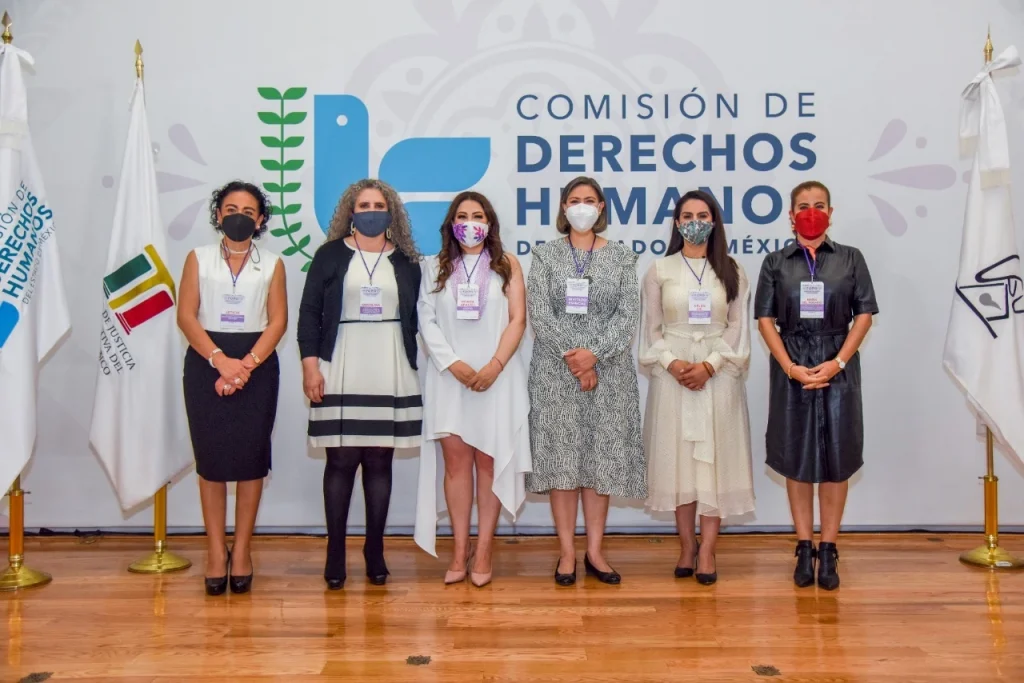 LAS MUJERES DEBEN CONSTRUIR REDES DE APOYO Y CONTRIBUIR A CREAR UNA SOCIEDAD MÁS DEMOCRÁTICA: MAGM. Imagen de un grupo de personas de pie en un acto institucional, frente al logotipo de la Comisión de Derechos Humanos del Estado de México.