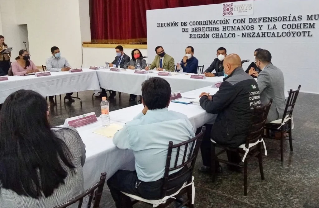 CONTINÚAN LAS REUNIONES DE COORDINACIÓN ENTRE LA CODHEM Y DEFENSORÍAS MUNICIPALES DE DDHH. Imagen de una mesa de trabajo con personas participantes en una sesión institucional, sentadas alrededor de mesas con documentos, en un salón oficial.