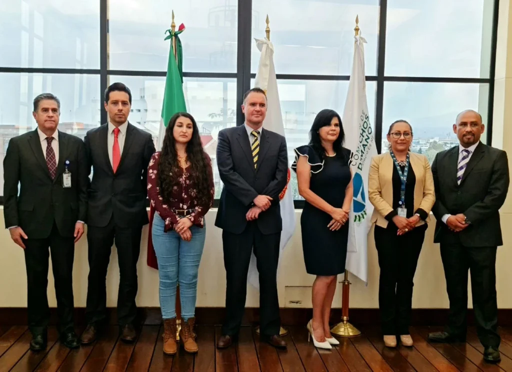 ESTRECHAN LAZOS DE COLABORACIÓN LA CODHEM Y EL MECANISMO PARA LA PROTECCIÓN INTEGRAL DE PERIODISTAS Y DEFENSORES DE DDHH. Imagen de un grupo de personas en un acto institucional, de pie frente a banderas y un fondo oficial, durante una reunión formal.