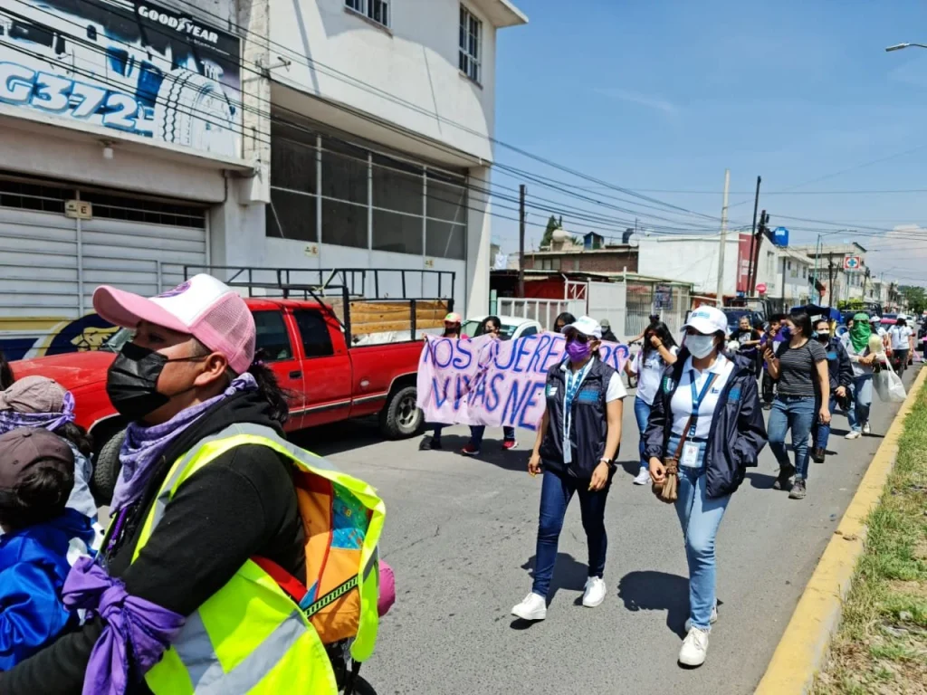 “Personas marchan por una calle con pancartas, mientras personal con chalecos institucionales acompaña el recorrido. La Codhem brindó acompañamiento durante la marcha para recordar a Diana Velázquez y corroboró que los elementos de seguridad pública respetaran el derecho humano a la manifestación.”
