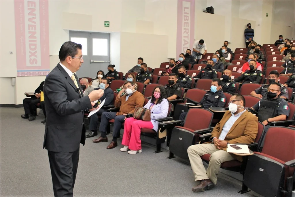 Persona de pie impartiendo una charla en un auditorio ante asistentes sentados, varios con uniforme policial y cubrebocas. Texto: “CAPACITA LA CODHEM A 100 POLICÍAS Y OFICIALES CALIFICADORES DE TOLUCA EN MATERIA DE DERECHOS HUMANOS”.