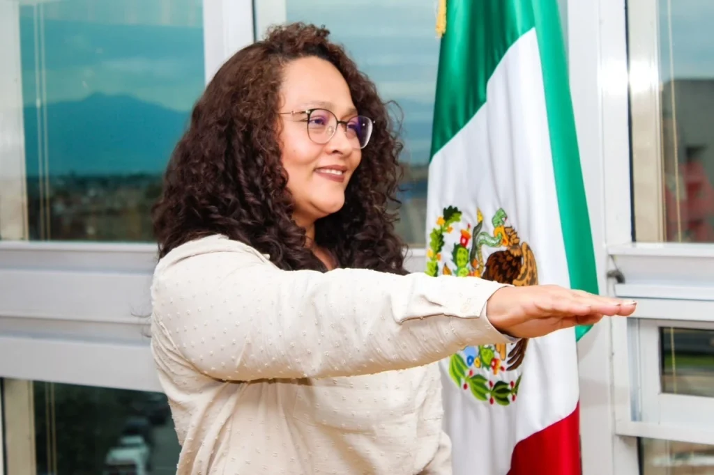 Persona levantando el brazo durante un acto protocolario frente a la bandera de México. Texto: “TOMA PROTESTA A NUEVA PRIMERA VISITADORA GENERAL LA PRESIDENTA DE LA CODHEM, Fabiola Manteca Hernández”.