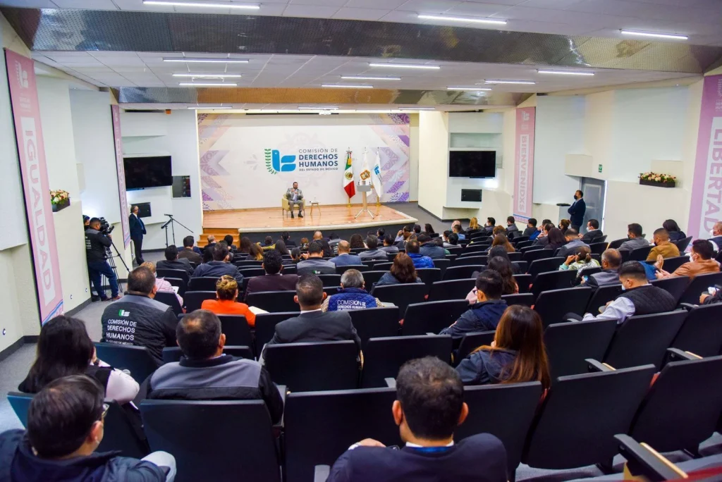 Auditorio con personal de la CODHEM sentado frente a un escenario durante una conferencia institucional. Texto: “PERSONAL DE LAS VISITADURÍAS DE LA CODHEM DEBE CONOCER EL SISTEMA PENAL ACUSATORIO Y SU MATIZ EN PROTECCIÓN DE DDHH: MAGM”.