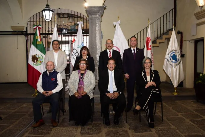 Grupo de personas posan para una fotografía institucional en un patio histórico, frente a banderas oficiales y emblemas de organismos públicos.