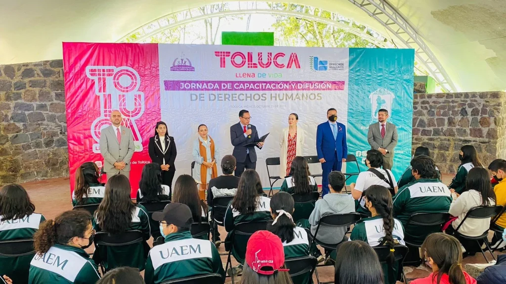 PROMUEVEN DERECHOS HUMANOS ENTRE ESTUDIANTES DE PREPARATORIA. Personas estudiantes sentadas frente a un escenario donde se realiza una jornada de capacitación sobre derechos humanos en Toluca.