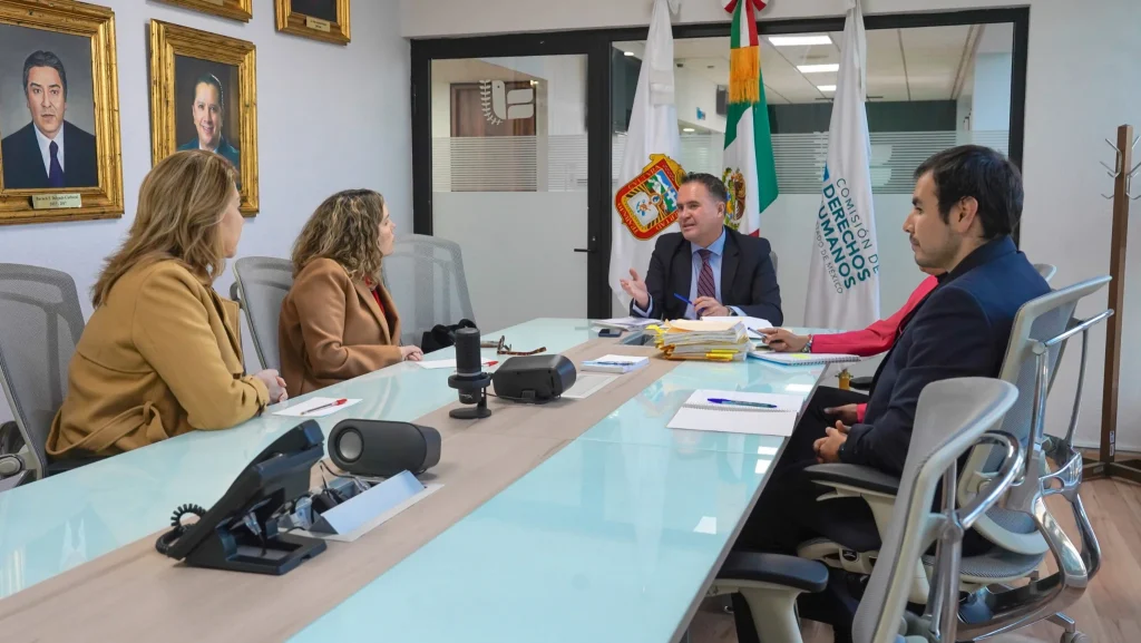 Reunión de trabajo en sala de juntas donde varias personas participan alrededor de una mesa con documentos, micrófonos y banderas institucionales al fondo; el encabezado “RESULTADOS DE LA CONSULTA INFANTIL Y JUVENIL 2021 DEBEN GENERAR POLÍTICAS PÚBLICAS PROTECTORAS DE LOS DDHH” resume el tema abordado.