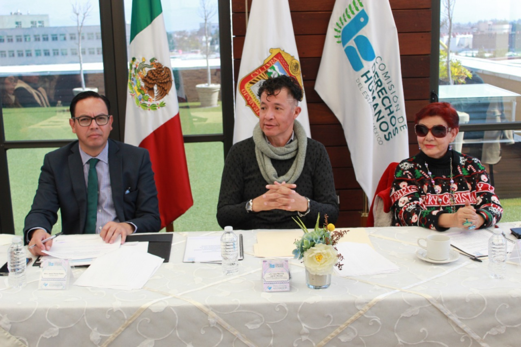 Tres personas en mesa de trabajo con documentos y banderas institucionales de fondo; el Consejo Ciudadano contra la Discriminación reafirma compromiso con grupos vulnerables y recibe gafetes de acreditación en la CODHEM.
