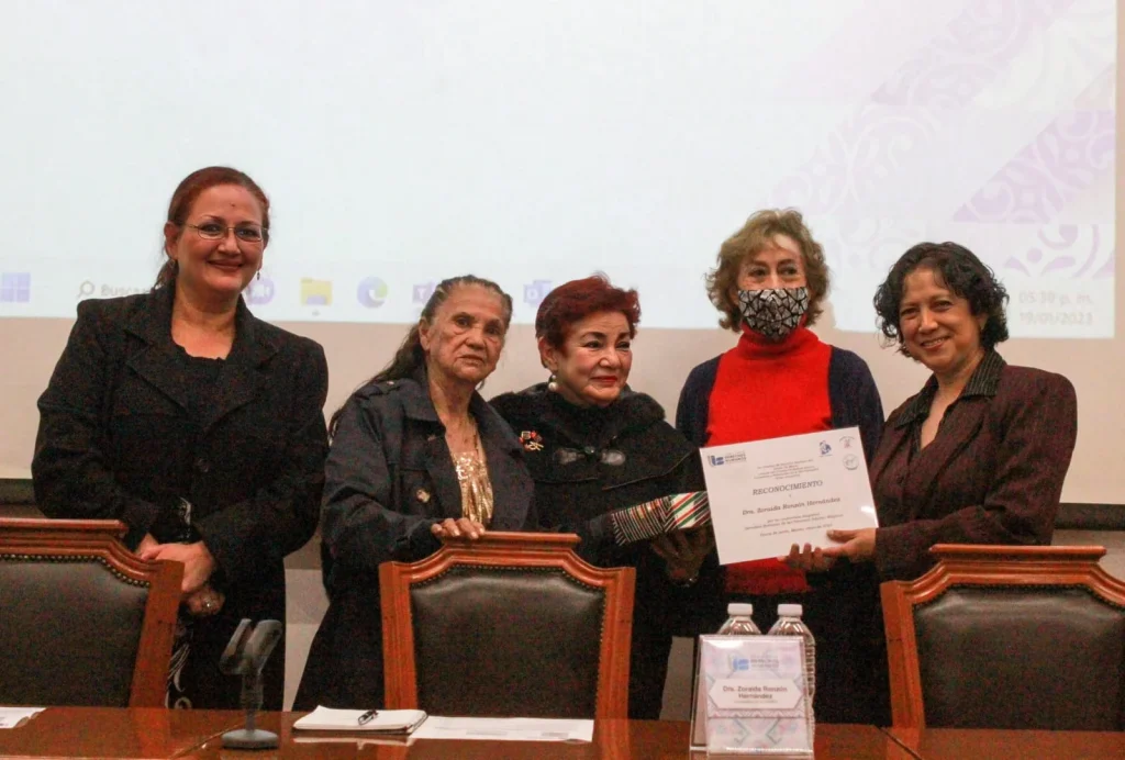 Cinco personas en evento institucional posan frente a mesa con micrófonos; una persona recibe y sostiene un reconocimiento impreso durante la ceremonia.