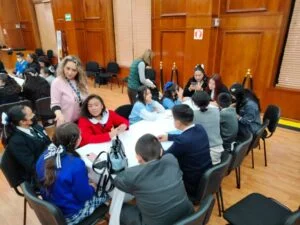 Grupo de personas reunidas alrededor de mesas en una sala con paredes de madera, participando en una dinámica de trabajo colaborativo con materiales y documentos sobre la mesa.