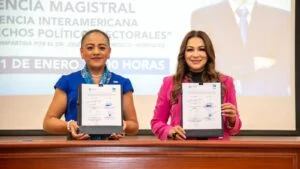 Dos personas en mesa de acto institucional sostienen reconocimientos ante un auditorio; se formaliza alianza estratégica entre instituciones para fortalecer derechos humanos y político-electorales.