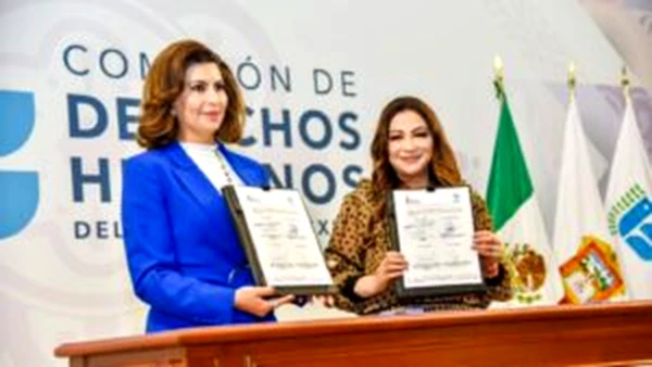 Dos personas sostienen documentos firmados durante un acto institucional frente a logotipos oficiales; se formaliza un convenio de colaboración y se presenta ponencia sobre derechos humanos laborales.