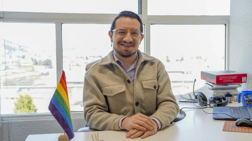 Persona servidora pública sentada en escritorio frente a una ventana amplia, con computadora portátil, audífonos y documentos; en primer plano destaca una bandera arcoíris, en un entorno de oficina institucional.