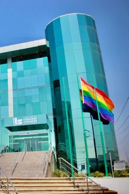 Edificio de la Comisión de Derechos Humanos del Estado de México con fachada de vidrio y bandera arcoíris al frente, en mensaje contra la homofobia.