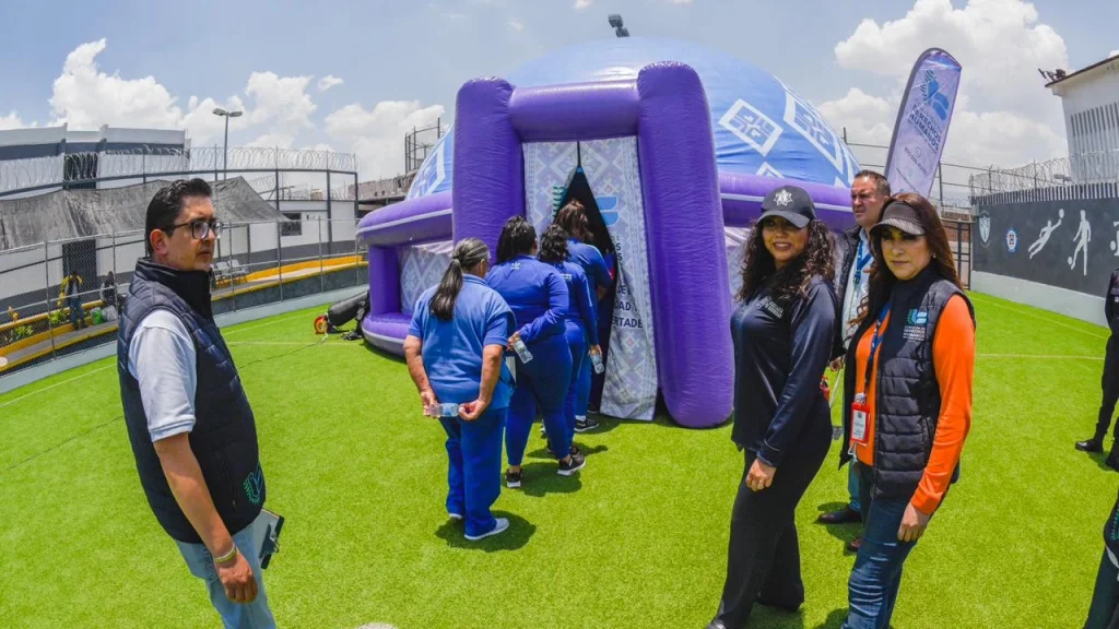 Fila de personas ingresando a una estructura inflable morada y azul instalada sobre un área de pasto, mientras otras personas observan alrededor en un espacio exterior cercado.