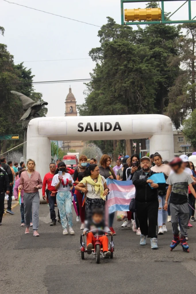 Grupo de personas participa en rodada y caminata por los derechos humanos en Toluca, avanzando por la calle bajo un arco de salida, con pancarta de diversidad visible.