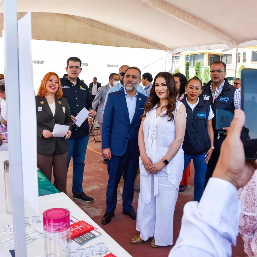 Personal de CODHEM y del INM recorre módulo informativo durante evento de atención a personas migrantes en el Estado de México, junto a materiales de orientación y registro.