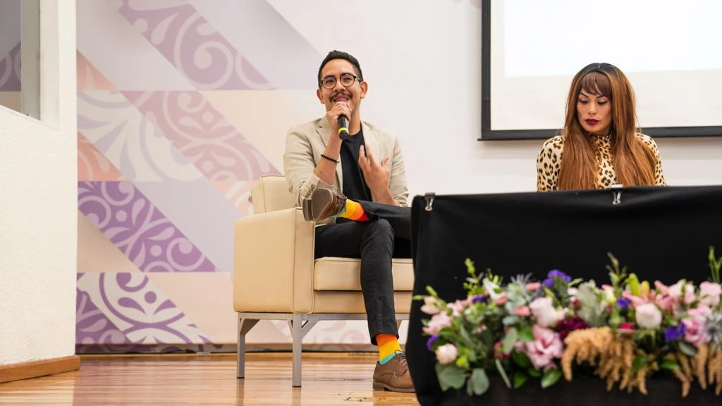 Persona sentada en un sillón hablando por micrófono durante un evento, acompañada por otra persona en el escenario; al frente hay una mesa con un arreglo floral.