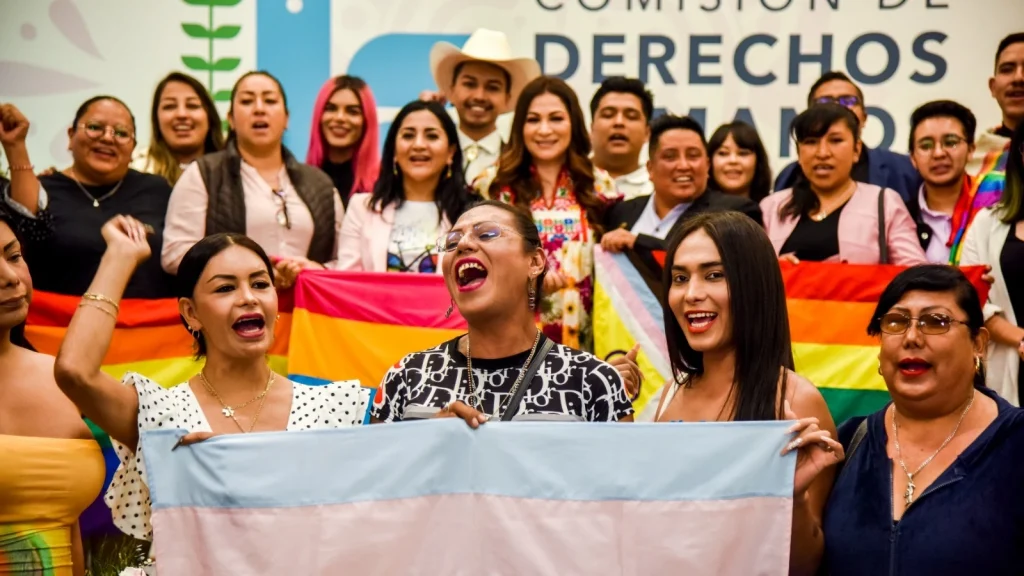Grupo de personas frente al logotipo de la CODHEM sostiene banderas de la diversidad y participa en una manifestación contra la discriminación institucional hacia la población LGBTTT+.