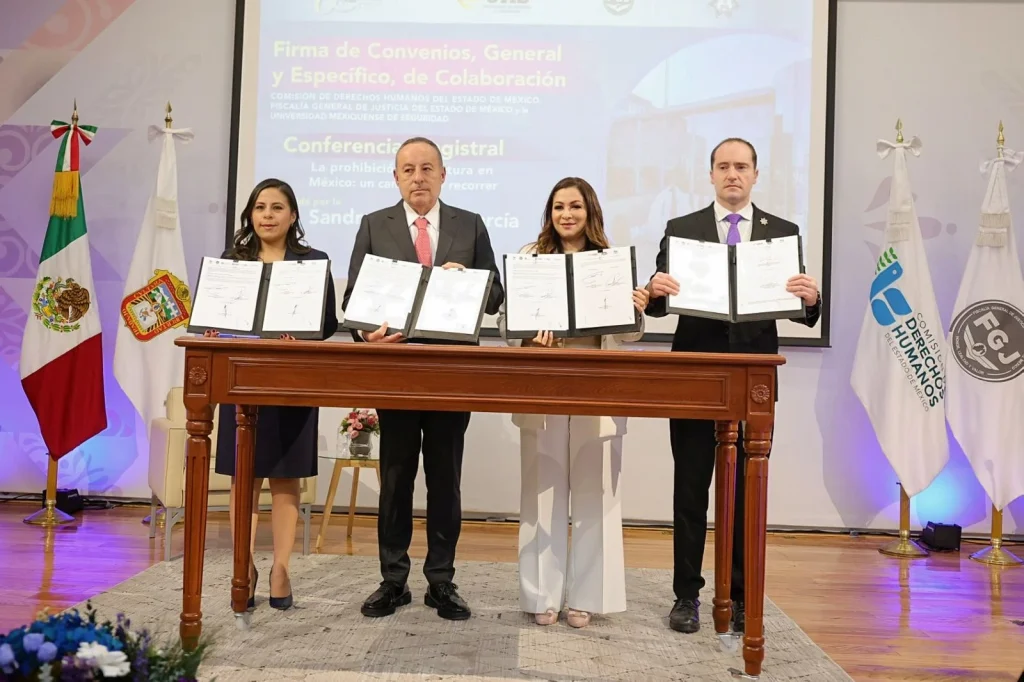Cuatro personas muestran documentos firmados durante la suscripción de convenios de colaboración entre la CODHEM, FGJEM, SSEM y la Universidad Mexiquense de Seguridad, frente a un fondo institucional con banderas oficiales.