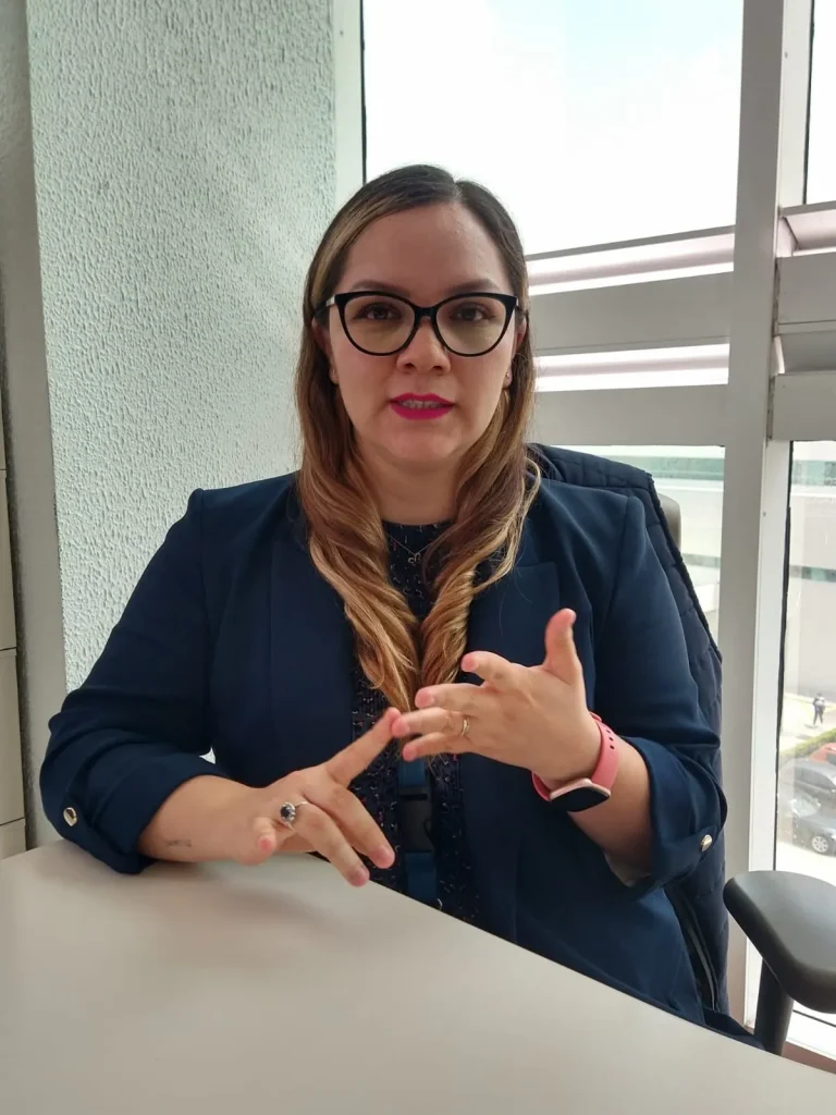 Persona con gafas sentada frente a un escritorio, realizando señas con las manos en un entorno de oficina, en alusión a la inclusión y los derechos de infancias con discapacidad.