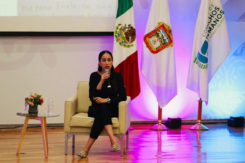 Persona participante sentada en escenario, hablando con micrófono durante un foro de la CODHEM, con banderas oficiales al fondo, en el contexto de políticas públicas para la primera infancia.