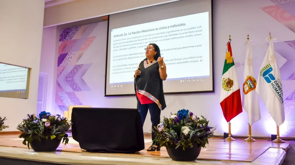 Persona expositora en escenario de la CODHEM presenta contenido sobre derechos de pueblos indígenas frente a una pantalla, con banderas oficiales al fondo, destacando la importancia de respetar su dignidad.Proporcione sus comentarios sobre BizChat