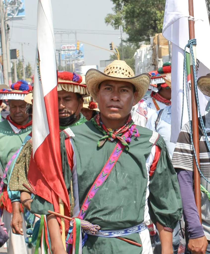 Grupo de personas con vestimenta tradicional participando en una marcha o evento público, portando banderas y atuendos típicos.