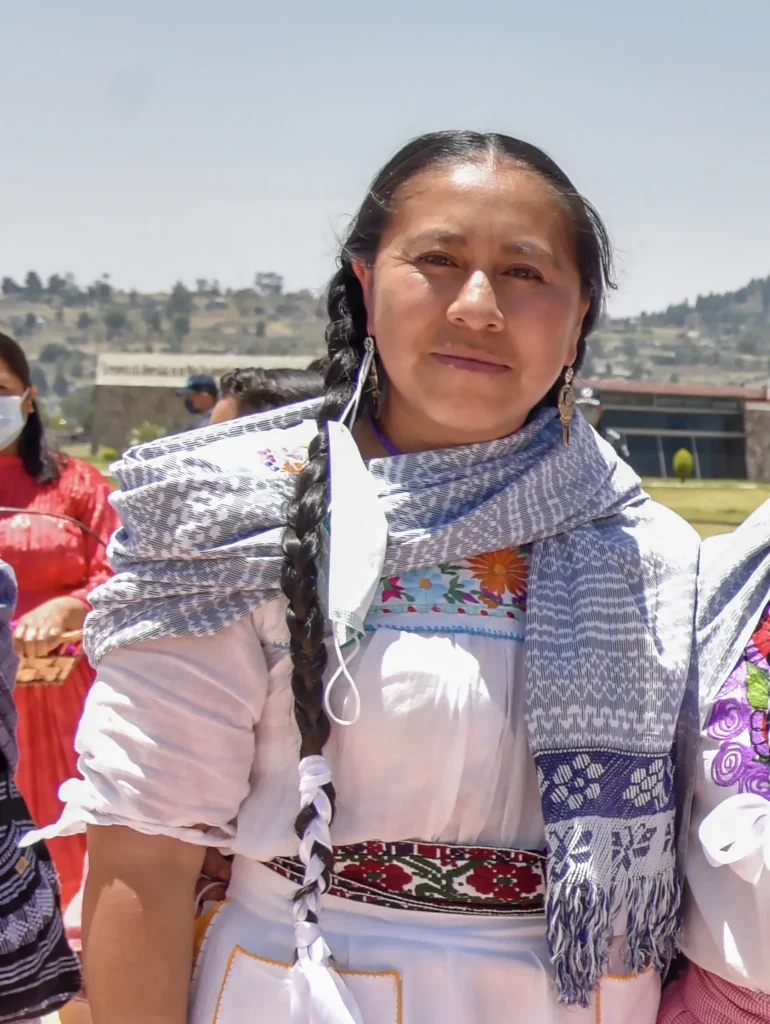 Persona con vestimenta tradicional y rebozo posando en un espacio abierto, con paisaje urbano al fondo y otras personas alrededor.