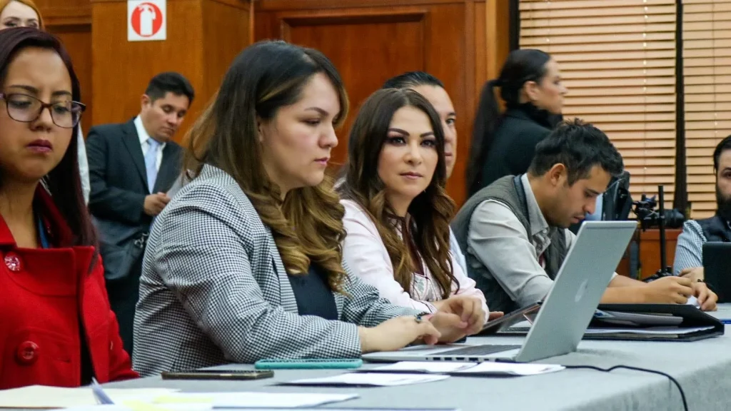 Personas participando en una reunión de trabajo, utilizando computadoras portátiles y revisando documentos en mesa de sesión.