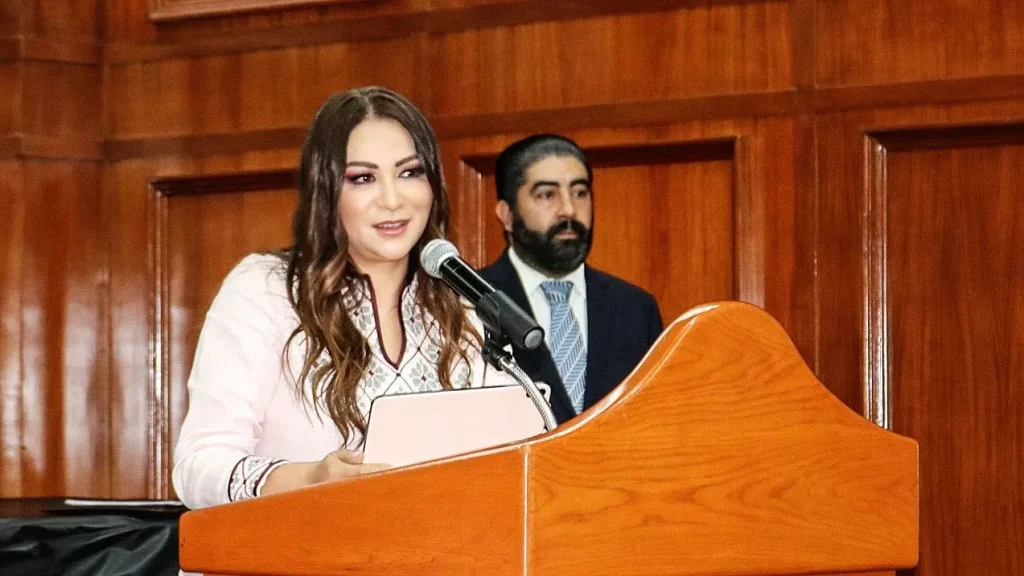 Persona hablando en un podio con micrófono, en un recinto con paredes de madera, durante una intervención pública.