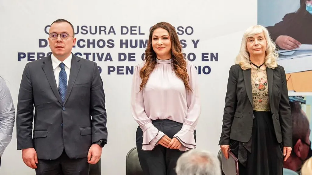 Tres personas de pie en un evento institucional frente a un fondo con la leyenda sobre la clausura de un curso de derechos humanos en el ámbito penitenciario, mientras público observa en primer plano.