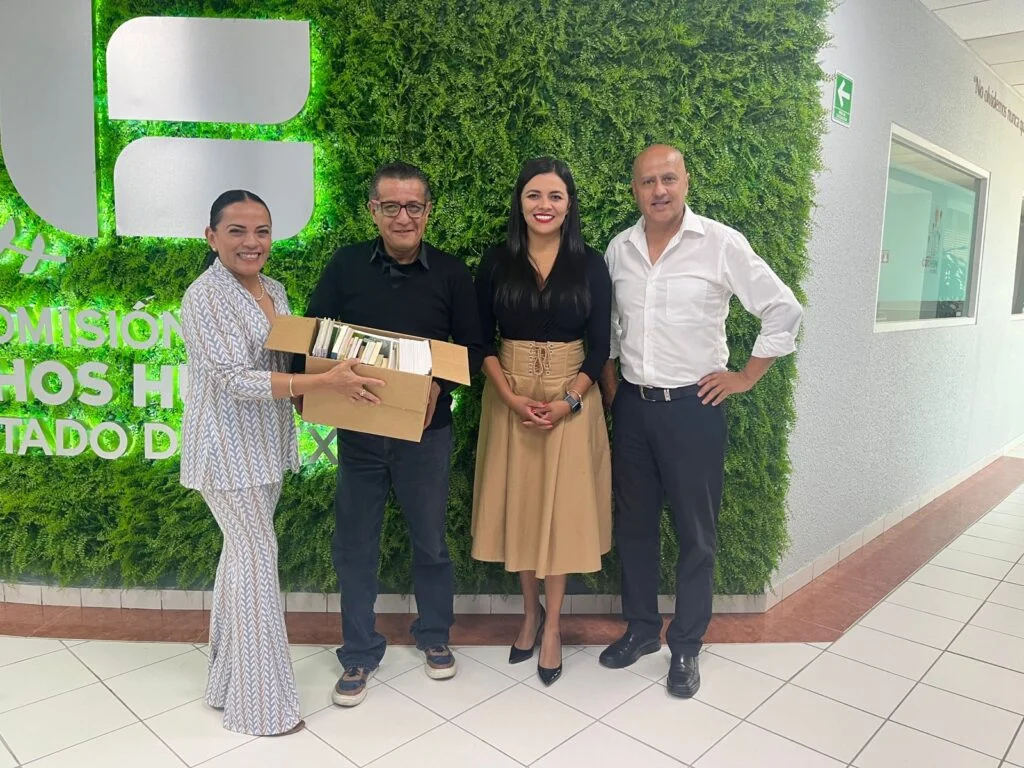 Cuatro personas posan frente a un muro verde con vegetación artificial en una oficina institucional; una de las personas sostiene una caja con libros, junto a un logotipo iluminado en la pared.