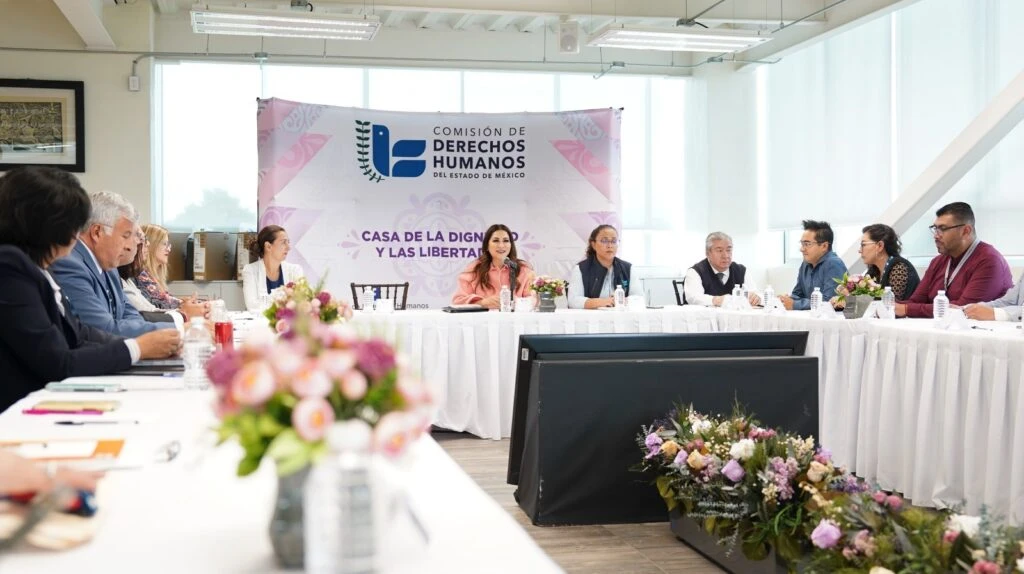 Reunión institucional en sala iluminada con varias personas sentadas alrededor de mesas blancas en forma de U, con arreglos florales al centro; al fondo se observa un banner que dice “Comisión de Derechos Humanos del Estado de México” y “Casa de la Dignidad y las Libertades”.