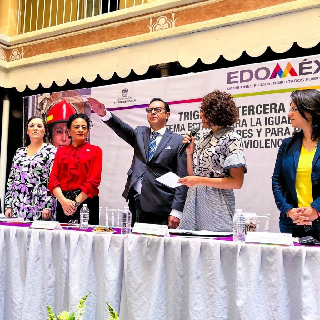 Personas de pie detrás de una mesa en un evento oficial frente a un banner del Gobierno del Estado de México (EDOMÉX); una persona al centro levanta la mano mientras otra sostiene un micrófono, en un acto público.