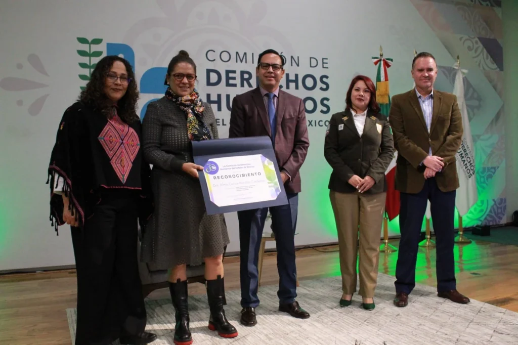Cinco personas posan en un escenario frente a un fondo con el logotipo de la Comisión de Derechos Humanos del Estado de México, mientras al centro se muestra un reconocimiento enmarcado durante un acto oficial.
