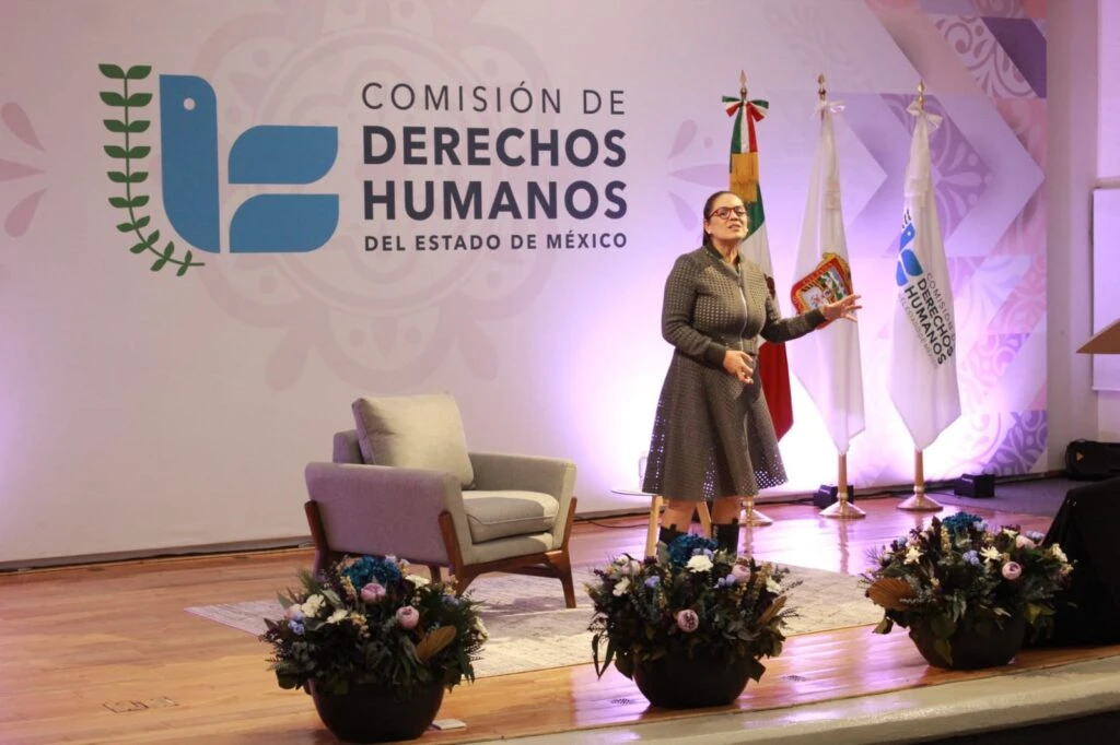 Persona en un escenario dando una ponencia frente a un fondo con el logotipo de la Comisión de Derechos Humanos del Estado de México, con una silla y arreglos florales al frente del área de exposición.