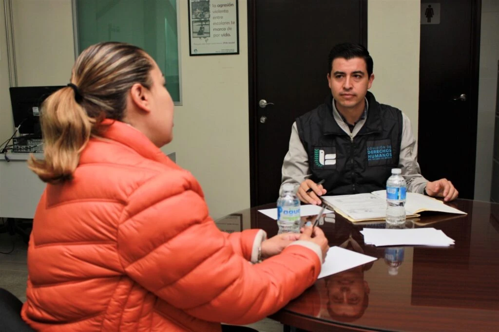 Dos personas sentadas frente a frente en una oficina sostienen una conversación en un escritorio con documentos y botellas de agua; una persona utiliza una libreta abierta mientras escucha.