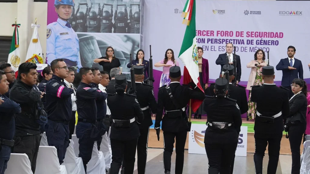Personal uniformado y asistentes participan en una ceremonia en interior; al centro se observa una bandera nacional y al fondo un escenario con un panel que menciona un “Foro de Seguridad con Perspectiva de Género” en el Estado de México.