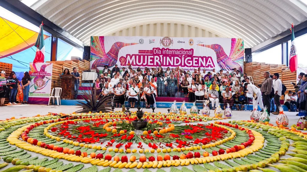Escenario con numerosas personas reunidas durante un evento del “Día Internacional de la Mujer Indígena”; al frente se observa una composición circular decorativa elaborada con frutas, hojas y flores, mientras al fondo hay un templete con un gran cartel colorido y banderas.