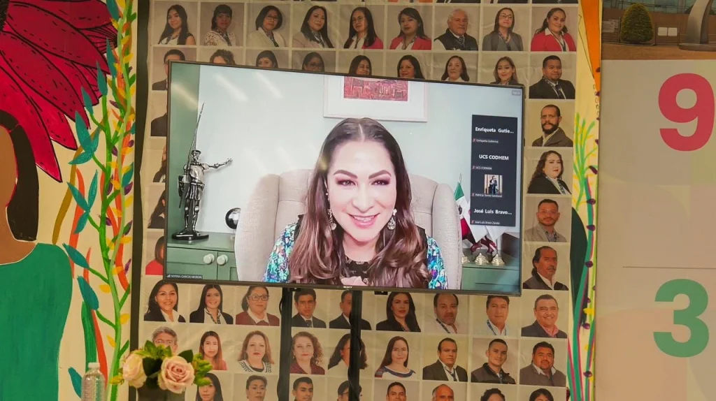 Pantalla de televisión mostrando a una persona participando en videoconferencia, colocada frente a un fondo con fotografías de múltiples retratos institucionales.