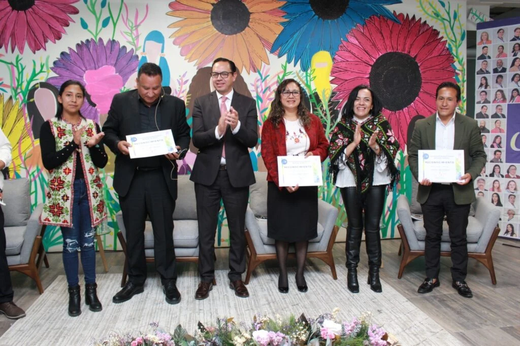 Grupo de seis personas de pie al frente de un espacio interior sosteniendo certificados y aplaudiendo durante una ceremonia; detrás se observa un mural colorido con flores grandes y figuras estilizadas, además de sillones y decoración floral en primer plano.