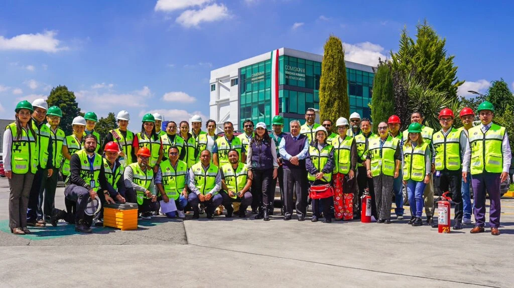 Grupo numeroso de personas posando al aire libre frente a un edificio institucional con fachada de vidrio; la mayoría porta cascos y chalecos de seguridad, y algunos sostienen equipo como extintores y maletines, en un contexto de actividad de protección civil bajo cielo despejado.